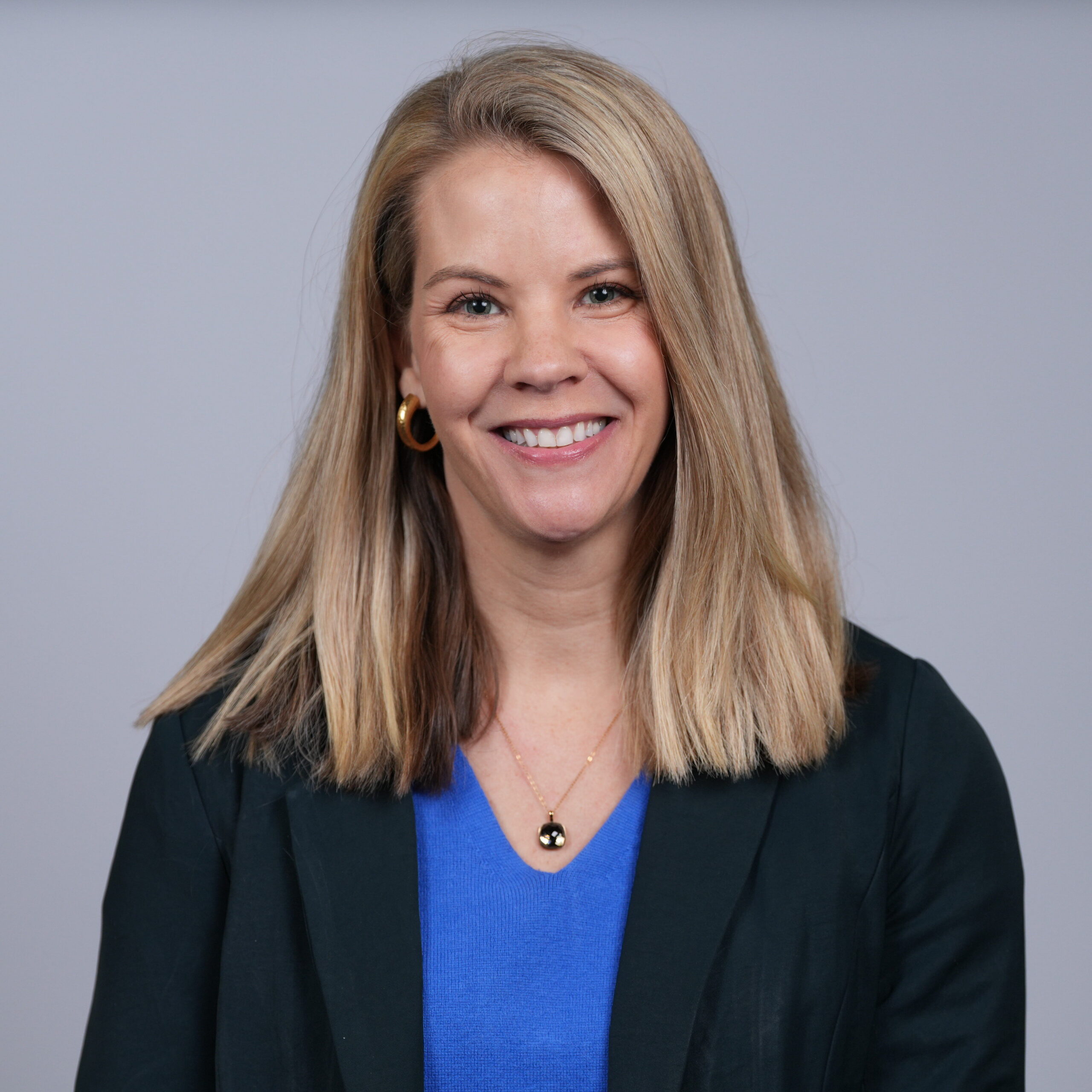 Close-up portrait of a smiling blonde woman wearing a blue top and dark blazer against a gray backdrop.